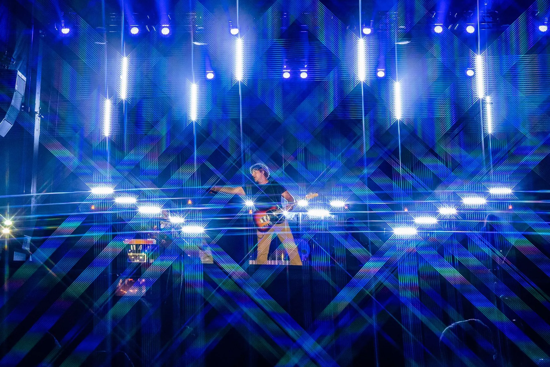 A guitarist on a stage framed by blue LED pillars and crisscrossing light streaks