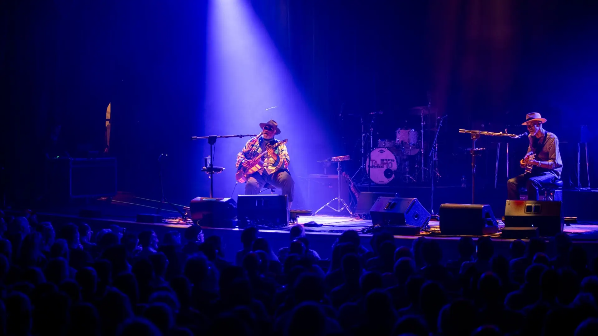 Two seated guitarists on a concert stage, one lit by a single violet spotlight beam through haze