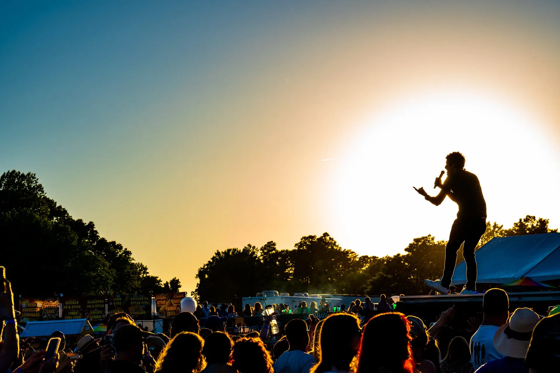 A singer silhouetted against the setting sun on an outdoor festival stage above the crowd