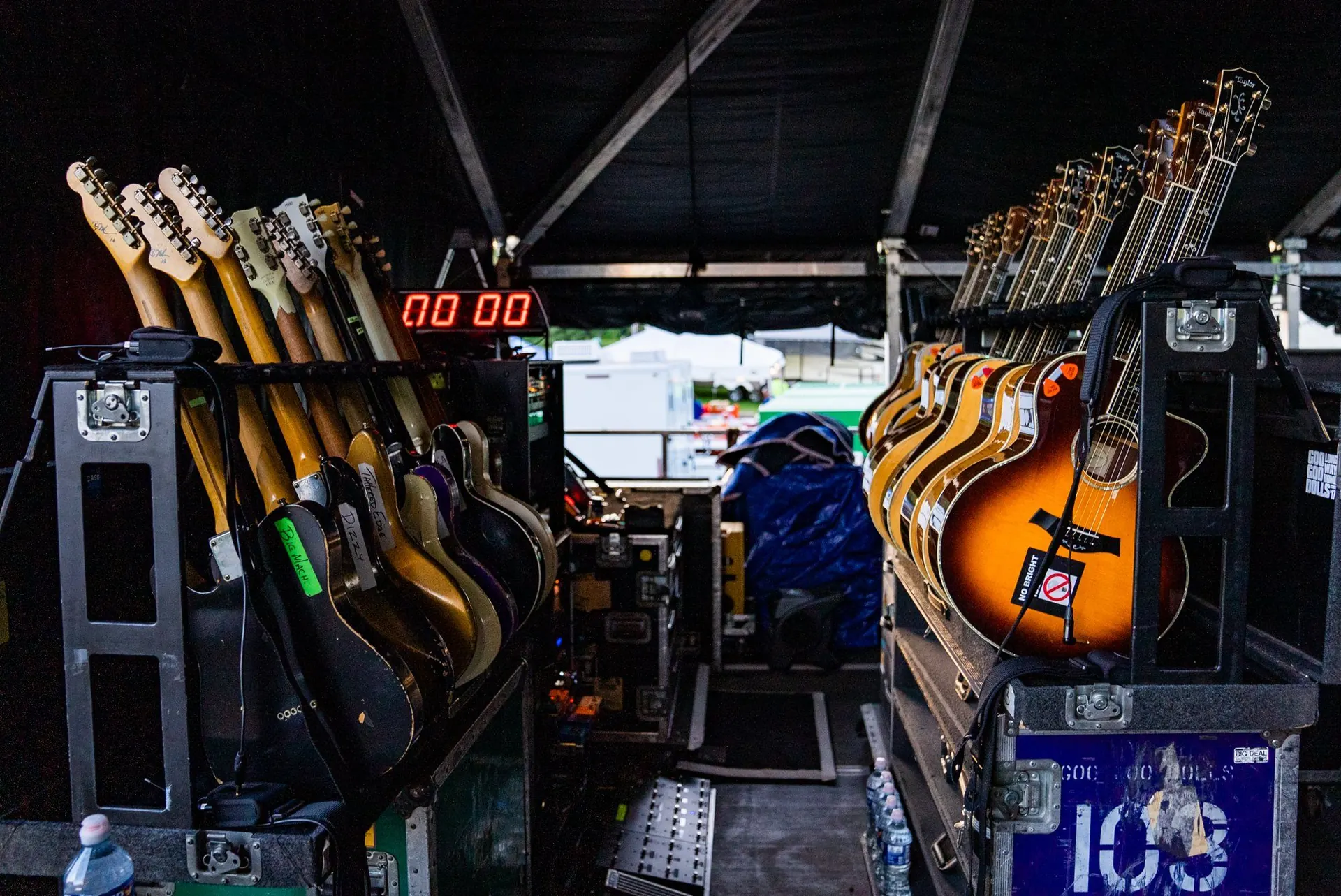 Backstage guitar world: racks of labeled electric and acoustic guitars on road cases flanking a show clock