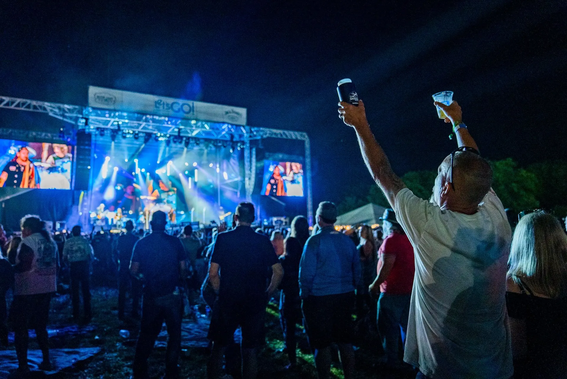 A fan raises two drinks toward a festival main stage with truss, LED screens and blue beams