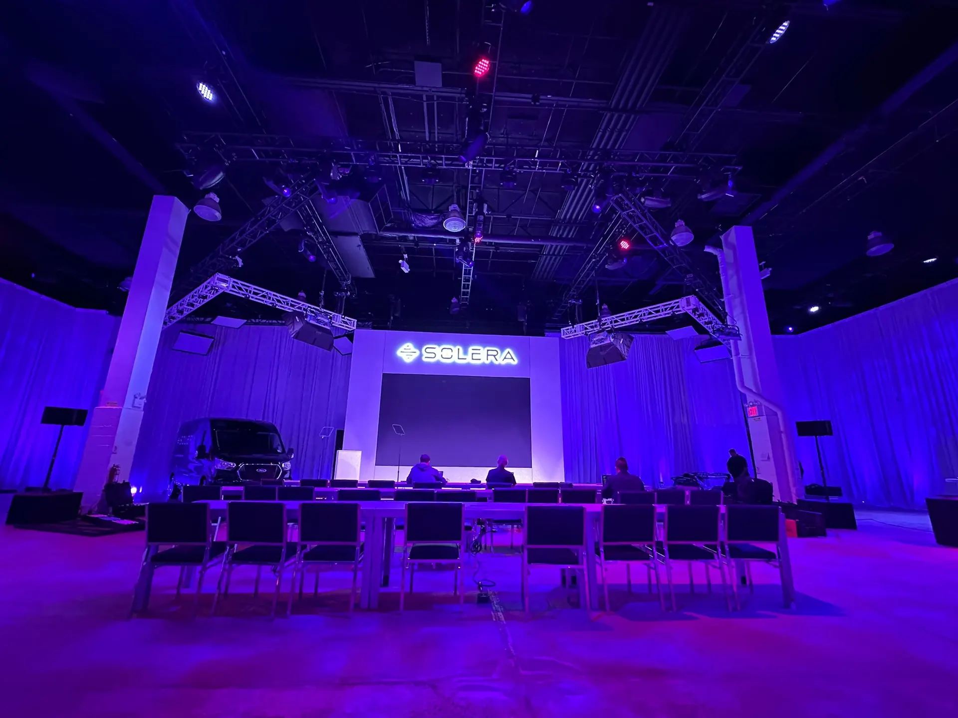 A corporate keynote set with an LED wall, lit signage, a vehicle display and rows of seating under a truss rig, washed in blue and violet light