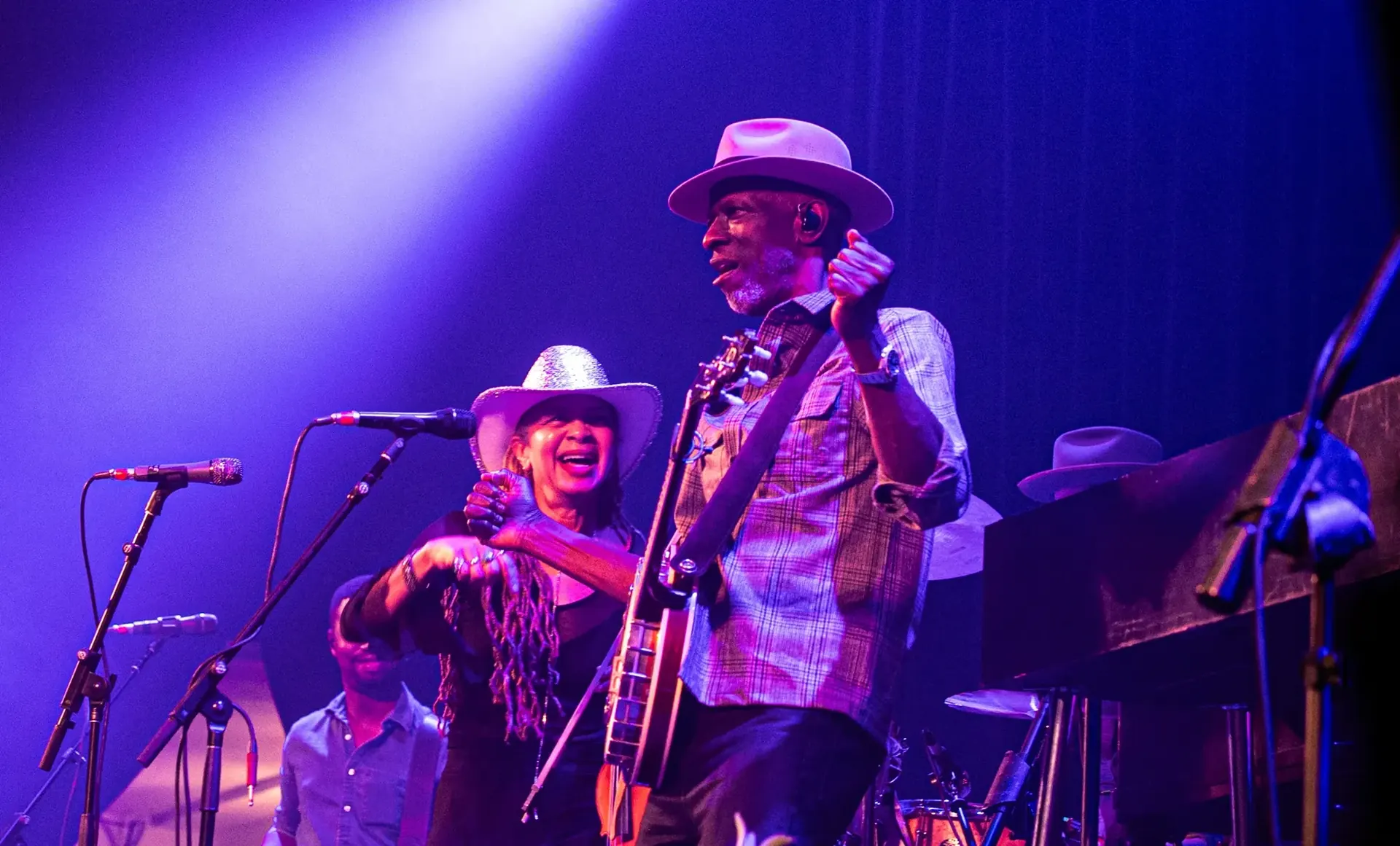Keb' Mo' with his banjo and a bandmate in a silver cowboy hat laughing on stage under violet light