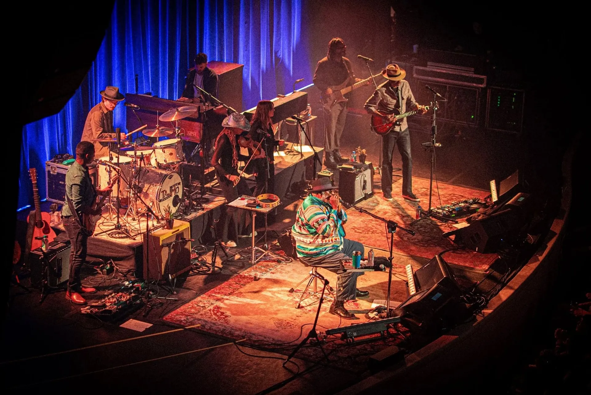 A full band on a rugged, multi-level stage with risers, backline and wedges, seen from the balcony